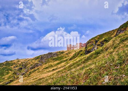 Una vista panoramica di una collina con lussureggiante erba verde e affioramenti rocciosi, caratterizzata da un edificio lontano arroccato sul pendio sotto un cielo parzialmente nuvoloso. Foto Stock