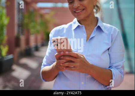 Una donna felice con una camicia blu che usa uno smartphone all'esterno. Sembra impegnata, mettendo in evidenza concetti di comunicazione, tecnologia e relax all'aperto Foto Stock
