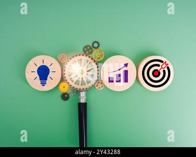 Icona target, simbolo della lampadina creativa e simbolo del grafico di crescita su blocchi di legno con ingranaggio in lente di ingrandimento su sfondo verde. Strato aziendale Foto Stock