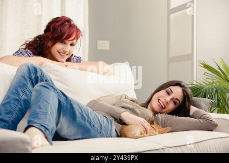 Due giovani donne allegre trascorrono del tempo insieme a casa, rilassandosi su un comodo divano con il loro piccolo cane. Stanno godendo di una spensierata e di gioia Foto Stock