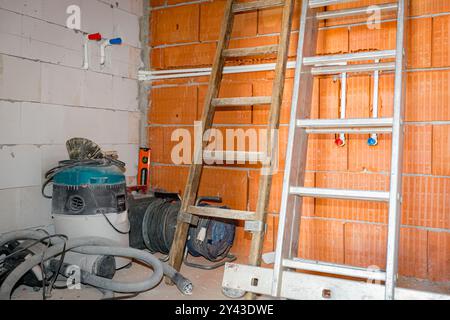 Scale in legno e metallo appoggiate contro pareti in blocchi rossi, collocate in un angolo del locale in costruzione, con aspirapolvere industriale e pochi aspirapolvere Foto Stock