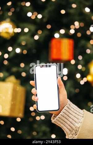 Primo piano di una mano che tiene uno smartphone davanti a un albero di Natale decorato con luci e ornamenti. Foto Stock