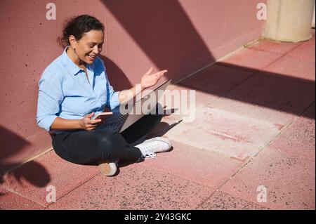 Una donna allegra siede su un marciapiede illuminato dal sole utilizzando un computer portatile e uno smartphone, godendosi una riunione virtuale all'aperto. Foto Stock