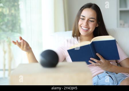 Donna felice che legge un libro di carta e parla con un altoparlante intelligente a casa Foto Stock