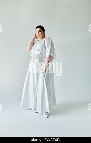 Una splendida sposa Plus size presenta il suo elegante abito bianco e gli orecchini scintillanti su uno sfondo grigio tenue. Foto Stock