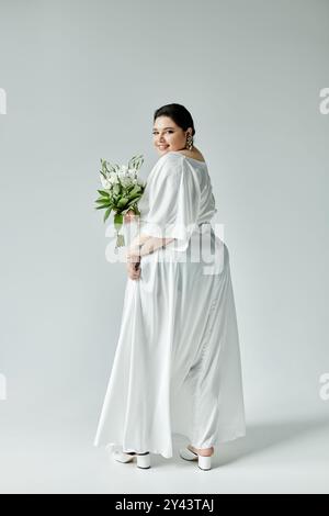 Una splendida sposa più grande mostra la sua eleganza in un abito bianco morbido, con un bellissimo bouquet. Foto Stock