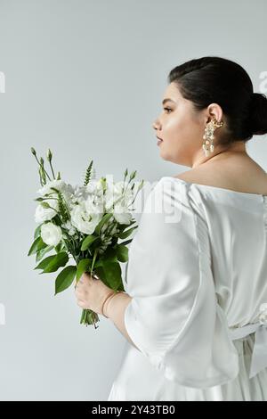 Una splendida sposa di grandi dimensioni mette in mostra il suo elegante abito da sposa e il suo bouquet su un morbido sfondo grigio. Foto Stock