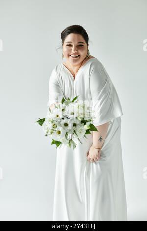 Una sposa radiosa e di grandi dimensioni irradia gioiosamente tenendo un bouquet su un elegante sfondo grigio. Foto Stock