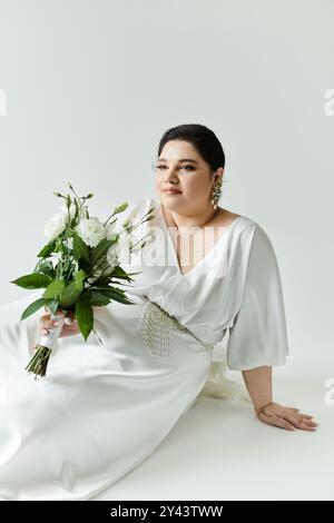 Una splendida sposa irradia bellezza in un abito bianco, che regge un bouquet lussureggiante, seduto graziosamente. Foto Stock