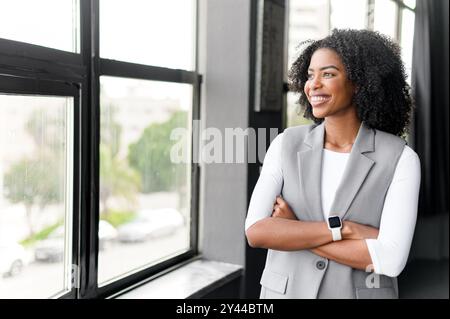 La donna d'affari sta in piedi in modo contemplativo vicino alla finestra, con le braccia incrociate, guardando verso la città, incarnando un senso di ambizione e di lungimiranza in un ambiente aziendale. Foto Stock