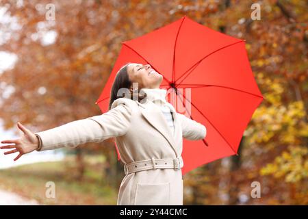 Donna felice di mezza età che respira ait fresco e che allunga le braccia festeggiando con un ombrello rosso in inverno in una foresta Foto Stock