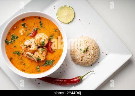 Tom Yam Kung zuppa tailandese piccante con gamberi e coriandolo in una ciotola bianca su sfondo chiaro con riso e pepe rosso, lime e bacchette su un piatto. Foto Stock