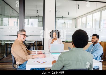 Un gruppo eterogeneo di professionisti collabora in un moderno ufficio, con una donna in un blazer bianco che conduce la discussione, indicativa di una sessione di brainstorming di squadra. Foto Stock