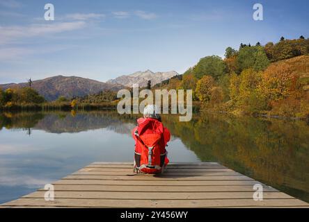 Una ragazza turistica con uno zaino sulla schiena ammira una fantastica vista delle montagne sedute sulla riva del lago Foto Stock