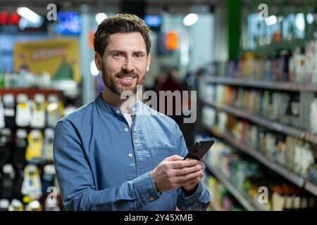 Uomo che tiene lo smartphone nella corsia del supermercato, sorride mentre è circondato da vari prodotti e scaffali. Cattura l'essenza della moderna esperienza di acquisto, dell'uso della tecnologia e della praticità nella vita quotidiana. Foto Stock