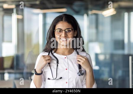 Ritratto di giovane ispanico medico donna all'interno dell'ufficio, operatore della clinica primo piano sorridente e guardando la macchina fotografica che indossa cappotto medico bianco con stetoscopio. Foto Stock