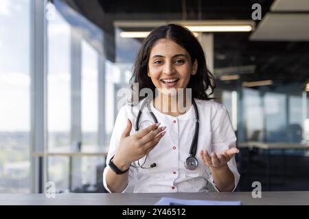 Visualizzazione videocamera Web, consulenza per videochiamate online, giovane medico che sorride e guarda la videocamera, comunica con la paziente da remoto, siede sul posto di lavoro all'interno della clinica, utilizzando un laptop. Foto Stock