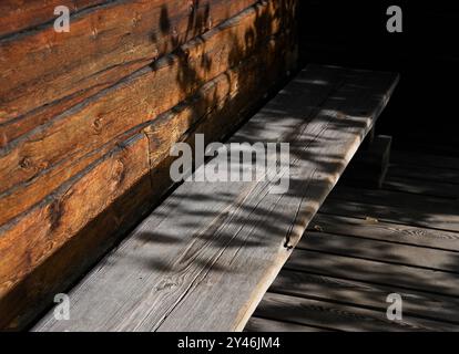 Questa immagine mostra una panchina rustica in legno all'ombra di una vecchia parete di legno. La luce naturale proietta ombre tenui delle piante o degli alberi vicini, crea Foto Stock