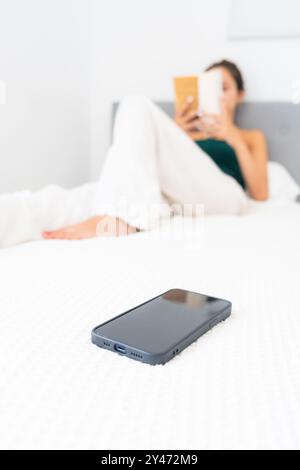 un adolescente che legge un libro con un telefono cellulare in primo piano si rilassa sul letto e si veste in modo informale. persone vere con luce naturale. Istruzione, re Foto Stock