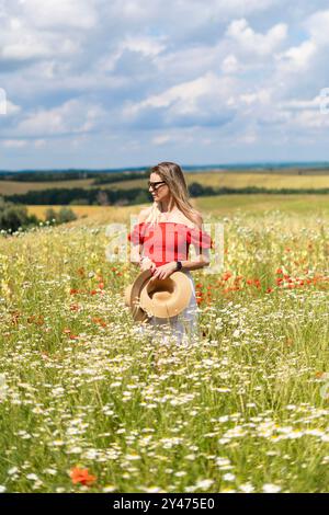 Donna bionda con occhiali da sole, vestito rosso e bianco in un campo di fiori selvatici, tenendo in mano cappelli di paglia Foto Stock