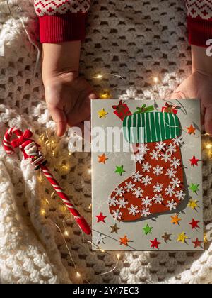 Calzino natalizio con biglietto d'auguri fatto a mano progetto fai da te. Carta colorata con borsa regalo di caramelle rosse per Babbo Natale. I bambini festeggiano le festività di Capodanno Foto Stock