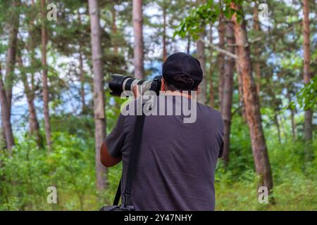 Un fotografo è adagiato tra alberi alti, utilizzando un grande obiettivo per catturare la fauna selvatica in una vivace foresta verde in una giornata limpida, circondato dalla bellezza della natura. Foto Stock