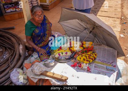 Mapusa, India - 19 febbraio 2024: Una vecchia donna seduta sul pavimento vende ghirlande di fiori. Mercato di strada Foto Stock