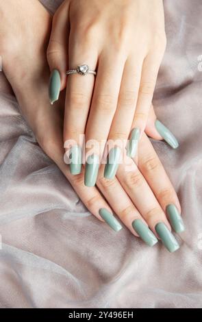 Un paio di mani con unghie lunghe e curate dipinte con una sfumatura di verde chiaro sono adagiate delicatamente su un morbido tessuto grigio chiaro. Foto Stock