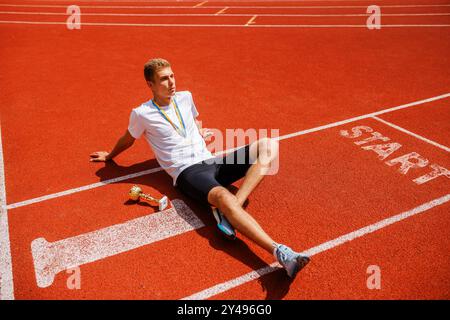 Un giovane atleta maschile seduto su una pista da corsa, sorridente e rilassato. Indossa una t-shirt bianca e pantaloncini neri, con una medaglia intorno al collo e. Foto Stock