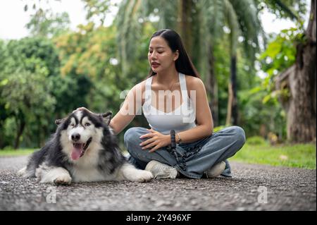 Un bellissimo husky siberiano al guinzaglio e la sua proprietaria asiatica sono seduti insieme su una strada in un parco verde, godendosi il loro tempo all'aperto. p Foto Stock