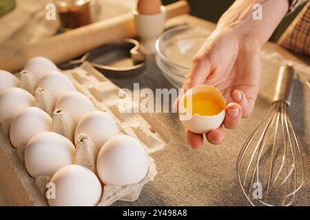Primo piano di mani femminili che tengono l'uovo di pollo rotto sullo sfondo del tavolo da cucina, cartone di uova, preparazione dell'impasto Foto Stock