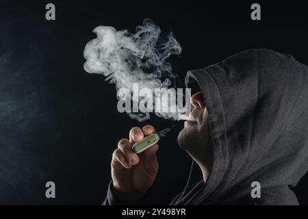 Uomo con mod vaping che espirano vapore su sfondo nero studio. Bearded Guy fuma sigaretta elettronica per smettere di fumare. SM libero da vapori e nicotina alternativa Foto Stock