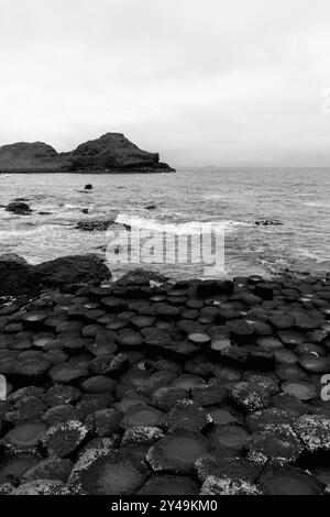 Foto monocromatica verticale del Selciato del Gigante, Irlanda del Nord, che mostra colonne esagonali di basalto che incontrano la costa atlantica, con una lontana scogliera rocciosa Foto Stock