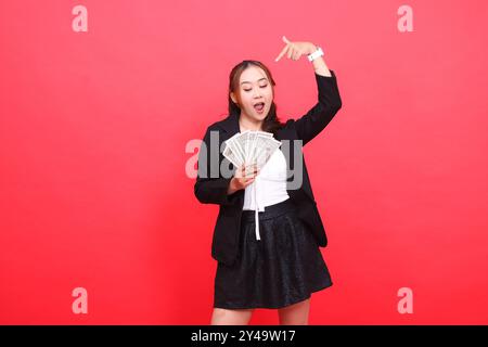 gesto della giovane donna d'ufficio indonesiana che guarda i soldi felice indossa un abito formale nero che tiene la banconota da un dollaro e lo indica dall'alto, per Foto Stock