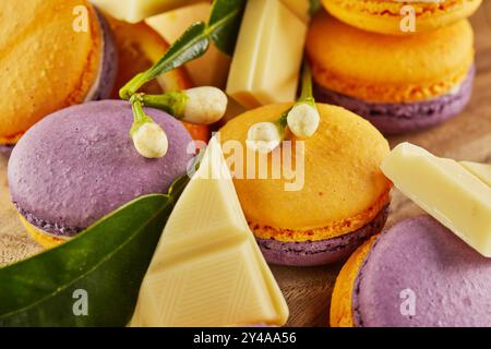 Vivaci macaron gialli e viola visualizzati con fette arancioni, foglie verdi e pezzi di cioccolato bianco su una tavola rustica di legno. Foto Stock