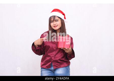 Foto ritratto di una bella ed elegante ragazza asiatica sui 20 anni che porta regali in scatola al cappello di Babbo Natale con una maglietta rossa moderna e lucida che mostra la g Foto Stock