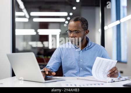 Uomo d'affari afroamericano maturo che lavora diligentemente sull'analisi finanziaria. Utilizza laptop e documenti stampati per rivedere i dati. Rappresenta il concetto di concentrazione, professionalità e tecnologia Foto Stock