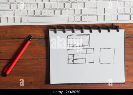 Un quaderno con disegni disegnati a mano, che include un layout con TV e mobili, collocato su un tavolo di legno accanto a una penna rossa e una tastiera per computer. Foto Stock