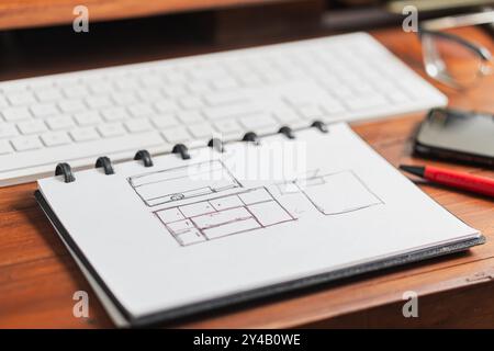 Un quaderno con disegni disegnati a mano, che include un layout con TV e mobili, collocato su un tavolo di legno accanto a una penna rossa e una tastiera per computer. Foto Stock