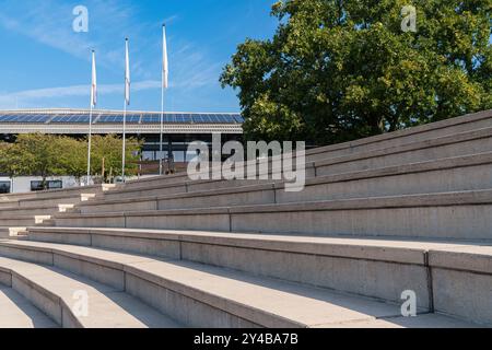 Una spaziosa area salotto in cemento simile a una scala si trova accanto ad un edificio contemporaneo. I pannelli solari sono visibili sul tetto, circondati da alberi e. Foto Stock