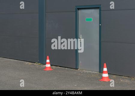 Una porta sicura si trova contro una parete testurizzata grigia, fiancheggiata da due coni stradali arancioni. L'impostazione suggerisce un'area ristretta con indicazione di attenzione Foto Stock