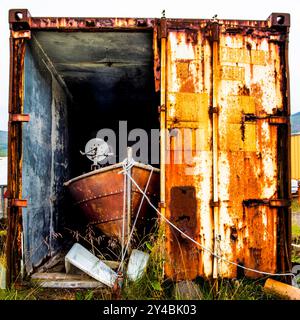 Piccola barca da pesca abbandonata all'interno di un container arrugginito con mezzo porta aperta nella pianura alla fine del fiordo a Borgarfiordur Eystri in Islanda Foto Stock