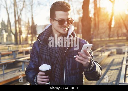 Ritratto di un uomo attraente che indossa una giacca, utilizza il telefono cellulare e tiene in mano il caffè da asporto mentre cammina per le strade della città Foto Stock