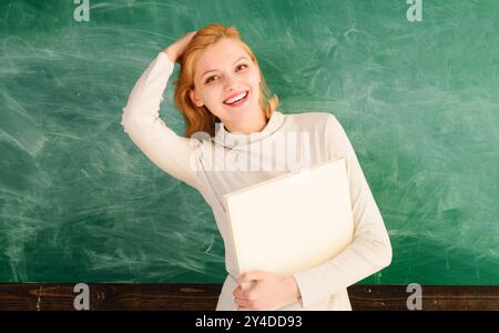 Insegnante o studentessa sorridente davanti alla lavagna con il libro della classe. Giornata degli insegnanti. Sorridente giovane studente o insegnante nel campus universitario con Foto Stock