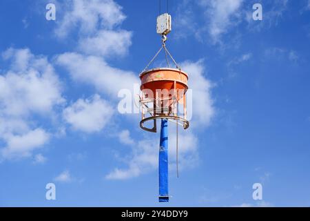 Un secchio arancione per calcestruzzo con tubo flessibile blu per il trasporto di calcestruzzo fresco è appeso a un gancio di gru in un cantiere edile contro un cielo blu Foto Stock