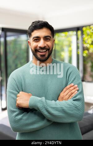 Uomo indiano sorridente con le braccia incrociate, indossa un maglione verde, in piedi all'interno Foto Stock