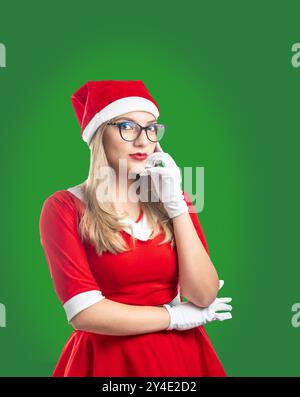 Giovane donna bionda con occhiali vestiti da Babbo Natale, che guarda seriamente la macchina fotografica su uno sfondo verde Foto Stock