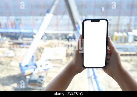 Primo piano di mani che tengono lo smartphone con schermo bianco vuoto su sfondo sfocato del cantiere Foto Stock