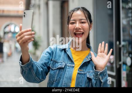 Una allegra donna asiatica con una giacca in denim sorride e saluta la mano alla telecamera, parla durante una videochiamata o registra il suo vlog di viaggio con una sma Foto Stock