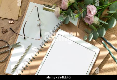 Uno spazio di lavoro luminoso è dotato di un tablet vuoto, un notebook aperto, utensili di cancelleria e fiori freschi disposti su un tavolo di legno, invitando la creatività e. Foto Stock